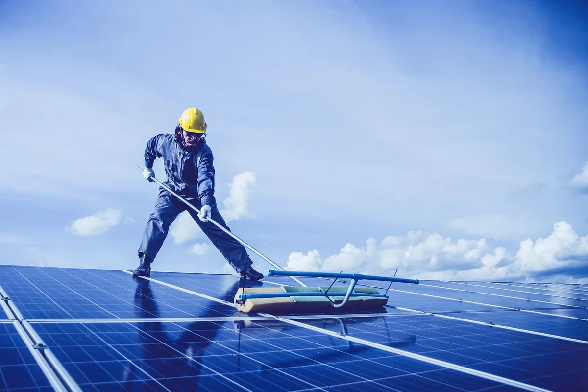 Operario con casco haciendo la limpieza de placas solares con un cepillo de mango largo