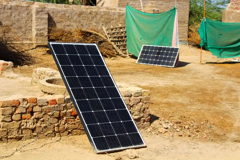 Placa solar apoyada en el brocal de ladrillo de un pozo, con su cable tendido por el suelo de tierra de una finca